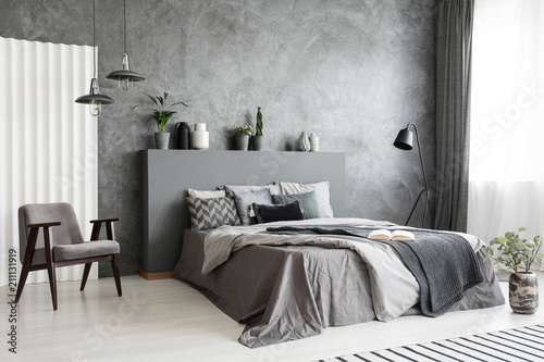 Modern grey bedroom interior with big bed with pillows and linen. Comfortable armchair next to the bed. Real photo.