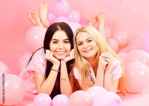 Gossip concept. Blonde and brunette on smiling faces have fun at domestic bedroom party. Girls lay on belly near balloons, pink background. Sisters or best friends in pajamas at girlish pajamas party.