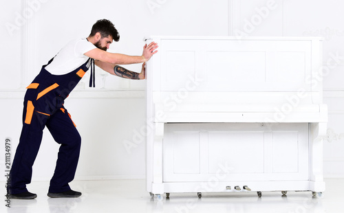 Man with beard and mustache, worker in overalls pushes piano, white background. Courier delivers furniture in case of move out, relocation. Courier service concept. Loader moves piano instrument. photo