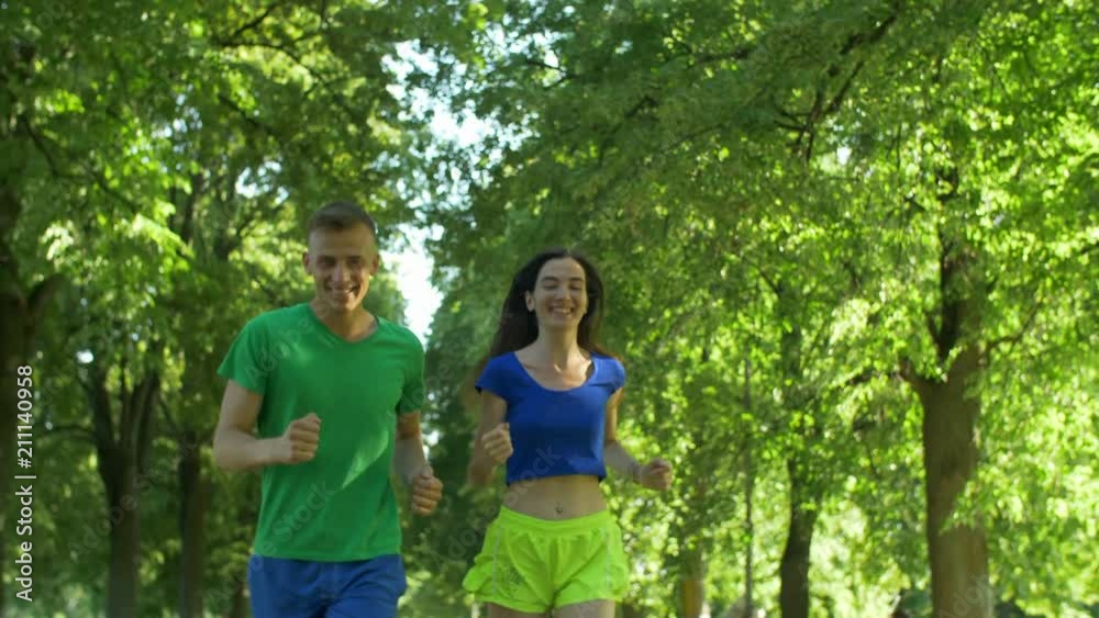 Portrait of cheerful sporty fit couple in sporswear exercising and jogging on park trail over beautiful landscape background. Positive athletic couple running together outdoors on sunny summer day. Portrait of cheerful sporty fit couple in sporswear exercising and jogging on park trail over beautiful landscape background. Positive athletic couple running together outdoors on sunny summer day.