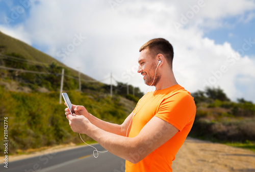 fitness, sport and technology concept - smiling young man with smartphone and earphones listening to music over big sur hills and road background in california