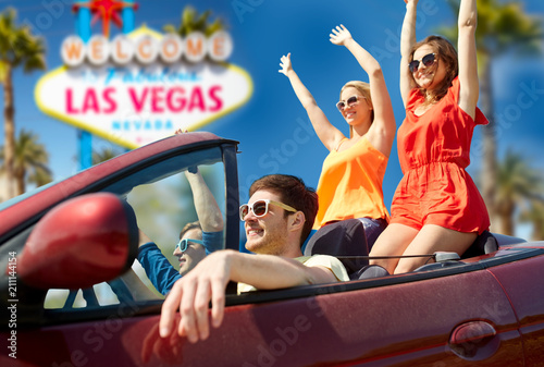 Fototapeta Naklejka Na Ścianę i Meble -  summer holidays, road trip and travel concept - happy friends driving in convertible car and waving hands over welcome to fabulous las vegas sign background