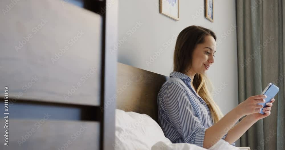 Young pretty woman sitting in her bed in the morning and using a tablet device, taping and scrolling. Inside