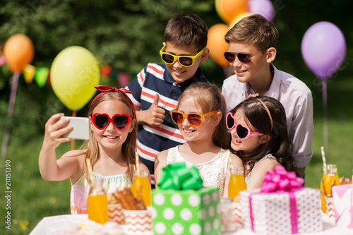 holidays, childhood and technology concept - happy kids in sunglasses taking selfie by smartphone on birthday party at summer garden