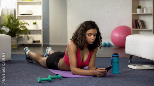 Fit young lady setting application on phone, lying mat, checking workout results