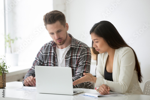 Thoughtful diverse colleagues working together on company business project, female Asian worker explaining issue to male coworker, focused employees analyzing financial statistics at laptop