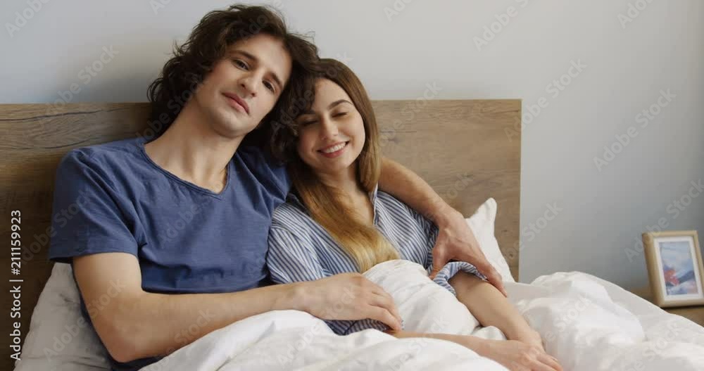 Portrait of the young good looking just-married man and woman in pajamas sitting in the bed and hugging in front of the camera. Indoors. Portrait of the young good looking just-married man and woman in pajamas sitting in the bed and hugging in front of the camera. Indoors.