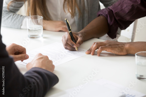 Close up of African American husband signing ownership contract buying first house with white wife, spouses closing deal with realtor or broker purchasing shared property or taking loan for apartment