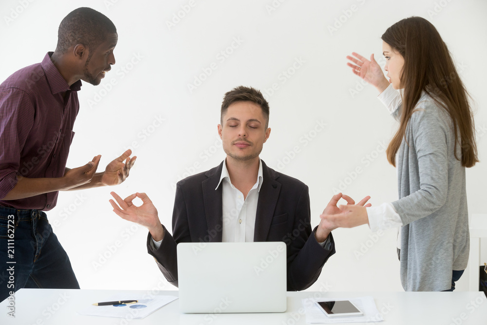 Caucasian male employee meditating, practicing yoga at workplace while multiracial colleagues yelling at him, blaming for business mistake or failure. Stress relief, emotion control, balance concept Caucasian male employee meditating, practicing yoga at workplace while multiracial colleagues yelling at him, blaming for business mistake or failure. Stress relief, emotion control, balance concept