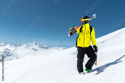 A portrait of a skier in a protective helmet and glasses is a mask and scarf with skis on his shoulder in the snow-capped mountains of the Caucasus. Skiing,