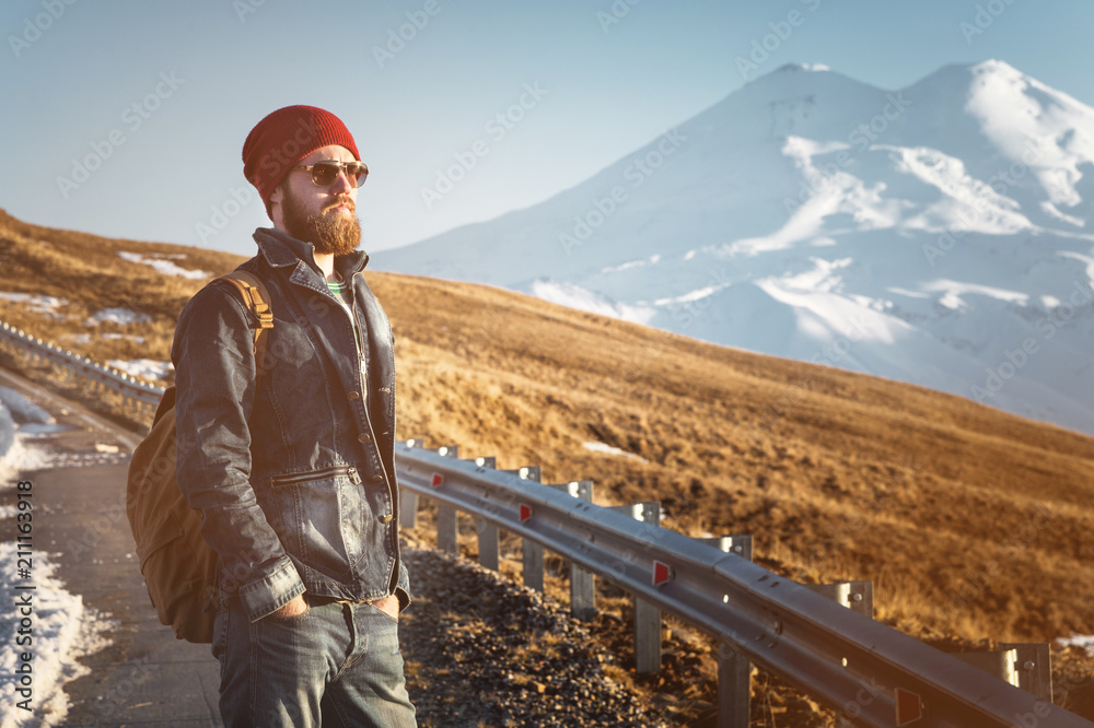 Bearded tourist hipster man in sunglasses with a backpack stand back on a roadside bump and watching the sunset against the background of a snow capped mountain Bearded tourist hipster man in sunglasses with a backpack stand back on a roadside bump and watching the sunset against the background of a snow capped mountain