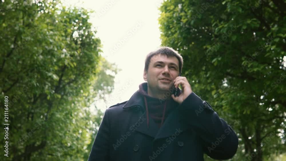 A young man in a black coat walks along the alley of blossoming apple trees in the park and talks on his mobile phone. A young man in a black coat walks along the alley of blossoming apple trees in the park and talks on his mobile phone.