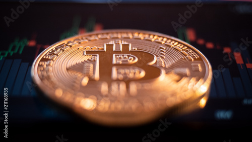 Close up of a golden glowing bitcoin. In the background, are candle stick graphs and courses on crypto stock exchanges. BTC coin is the symbol of decentralized cryptocurrency based on blockchain.