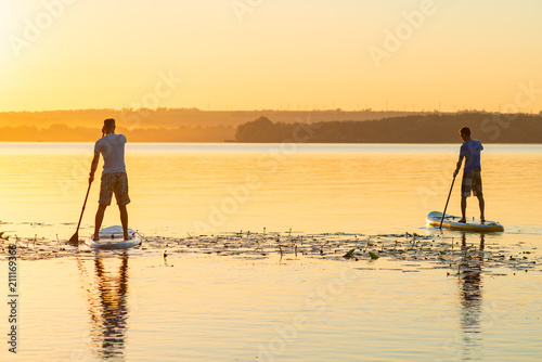 Men, friends sail on a SUP boards in a large river during sunrise