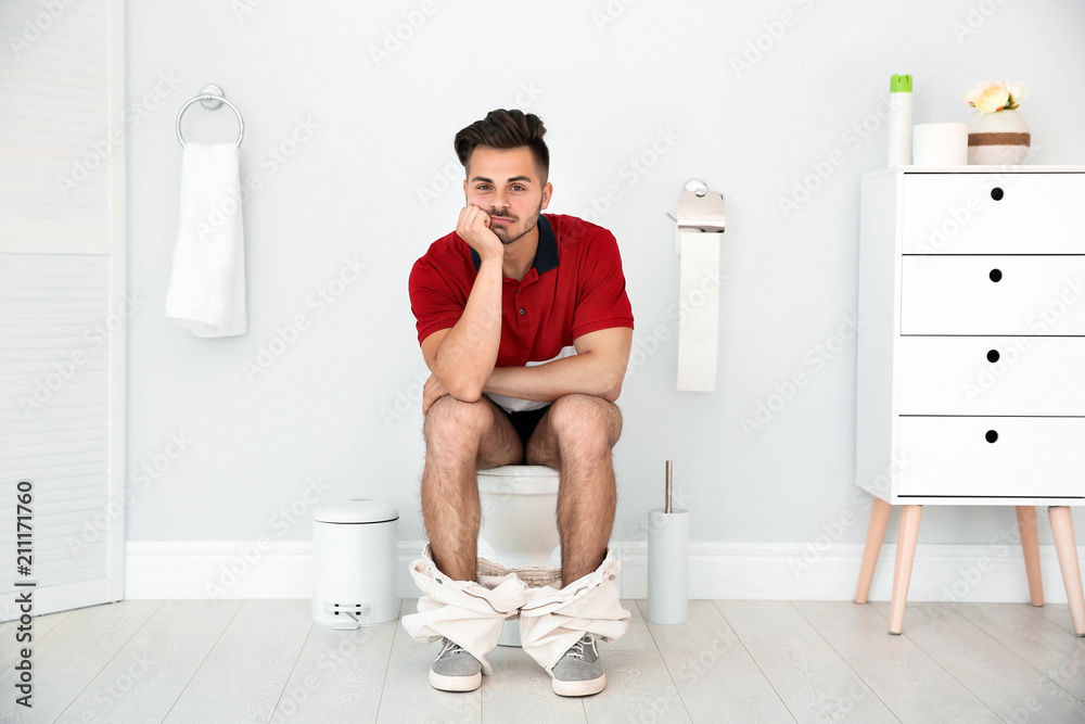 Young man sitting on toilet bowl at home - obrazy, fototapety, plakaty 