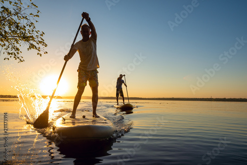Leinwand Poster Men, friends sail on a SUP boards in a rays of rising sun