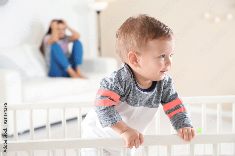 Cute baby boy in crib and young mother suffering from postnatal depression on blurred background Cute baby boy in crib and young mother suffering from postnatal depression on blurred background