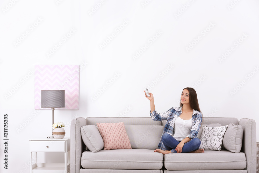 Young woman switching on air conditioner while sitting on sofa near white wall Young woman switching on air conditioner while sitting on sofa near white wall
