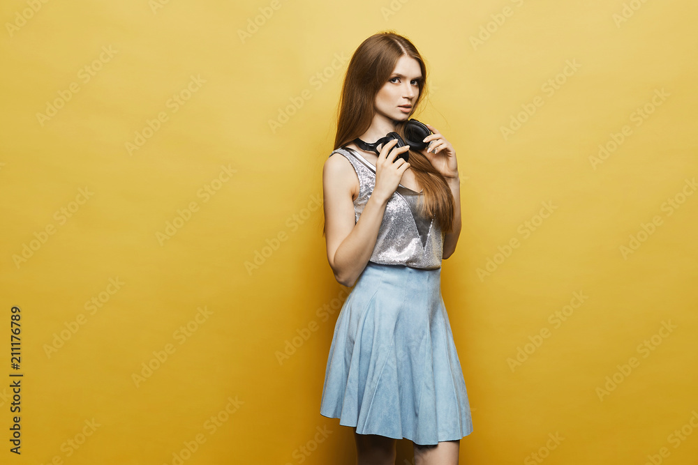 Sexy and beautiful blonde model girl with gentle makeup and with headphones on her neck, in fashionable skirt and in stylish silver blouse posing at yellow background, isolated Sexy and beautiful blonde model girl with gentle makeup and with headphones on her neck, in fashionable skirt and in stylish silver blouse posing at yellow background, isolated