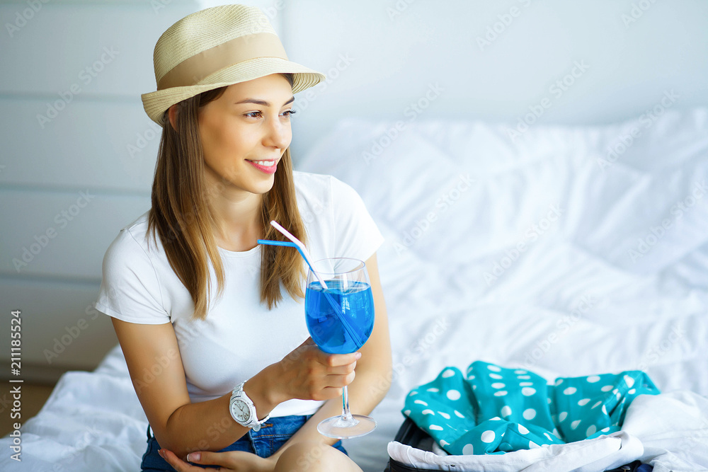 Vacation. Young Beautiful Girl Sits on the Bed Dressed in a Hat and Holding in Hands Blue Cocktail. Happy Girl Goes On Vacation. Portrait of the Woman who is Watching in the Window Vacation. Young Beautiful Girl Sits on the Bed Dressed in a Hat and Holding in Hands Blue Cocktail. Happy Girl Goes On Vacation. Portrait of the Woman who is Watching in the Window