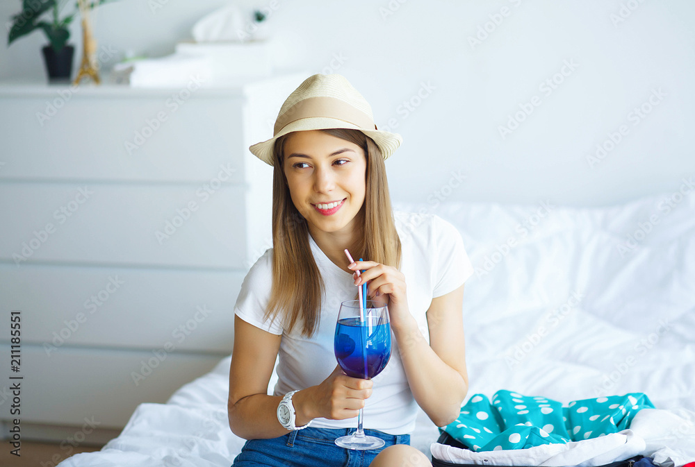 Vacation. Young Beautiful Girl Sits on the Bed Dressed in a Hat and Holds Cocktail in Hands. Happy Girl Goes On Vacation. High Resolution Vacation. Young Beautiful Girl Sits on the Bed Dressed in a Hat and Holds Cocktail in Hands. Happy Girl Goes On Vacation. High Resolution