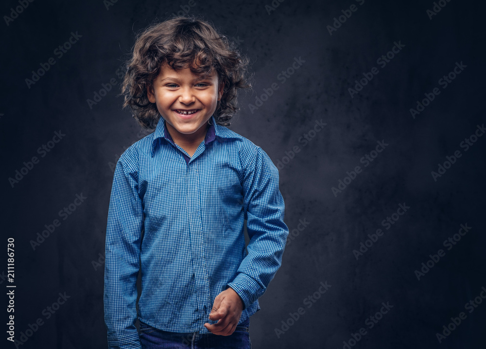 Cute smiling schoolboy with brown curly hair dressed in a blue shirt, posing at a studio. Isolated on the dark textured background. Cute smiling schoolboy with brown curly hair dressed in a blue shirt, posing at a studio. Isolated on the dark textured background.