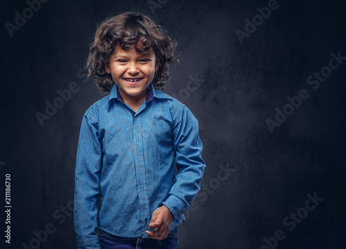 Cute smiling schoolboy with brown curly hair dressed in a blue shirt, posing at a studio. Isolated on the dark textured background.