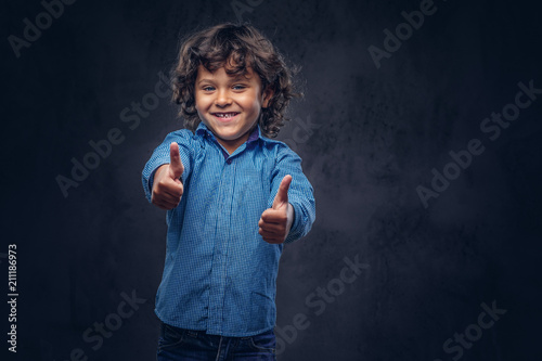 Contented boy cute schoolboy with brown curly hair dressed in a blue shirt, showing thumbs up at the studio.