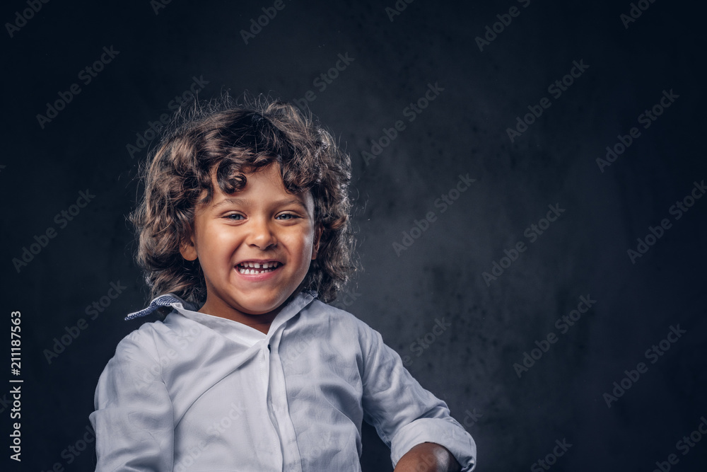 Cute smiling schoolboy with brown curly hair dressed in a white shirt, posing at a studio. Isolated on the dark textured background. Cute smiling schoolboy with brown curly hair dressed in a white shirt, posing at a studio. Isolated on the dark textured background.