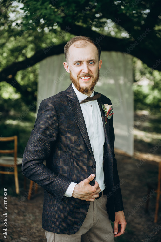 The groom. Portrait of a handsome smiling groom with a beard, with blue eyes, in a stylish suit. On nature The groom. Portrait of a handsome smiling groom with a beard, with blue eyes, in a stylish suit. On nature