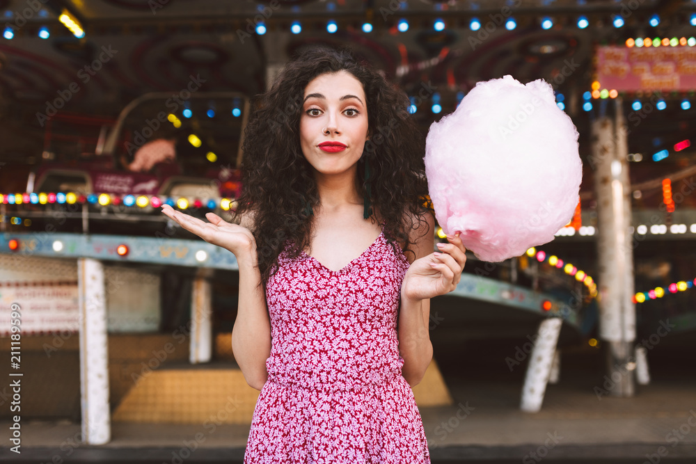 Young woman with dark curly hair in dress standing with cotton candy in  hand and thoughtfully looking in camera while spending time in amusement  park Stock Photo | Adobe Stock