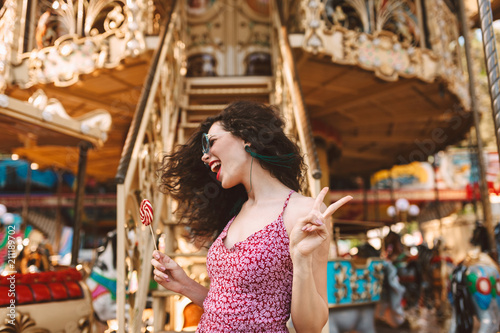 Cheerful woman with dark curly hair in sunglasses and dress standing with lolly pop candy in hand and happily looking aside while spending time in amusement park