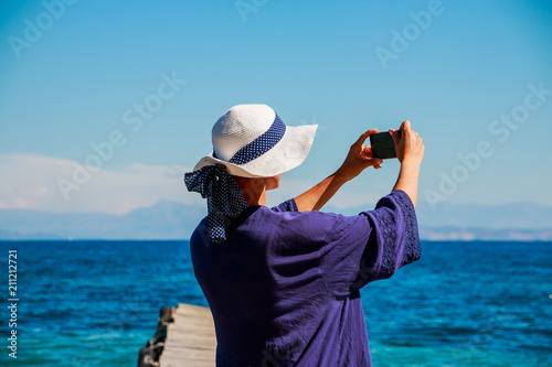 Beautiful girl in white summer hat and blue shirt on the beach makes selfie, photo. Stories from vacation for internet. Summer holiday on sea. Sunny weather. Copy space