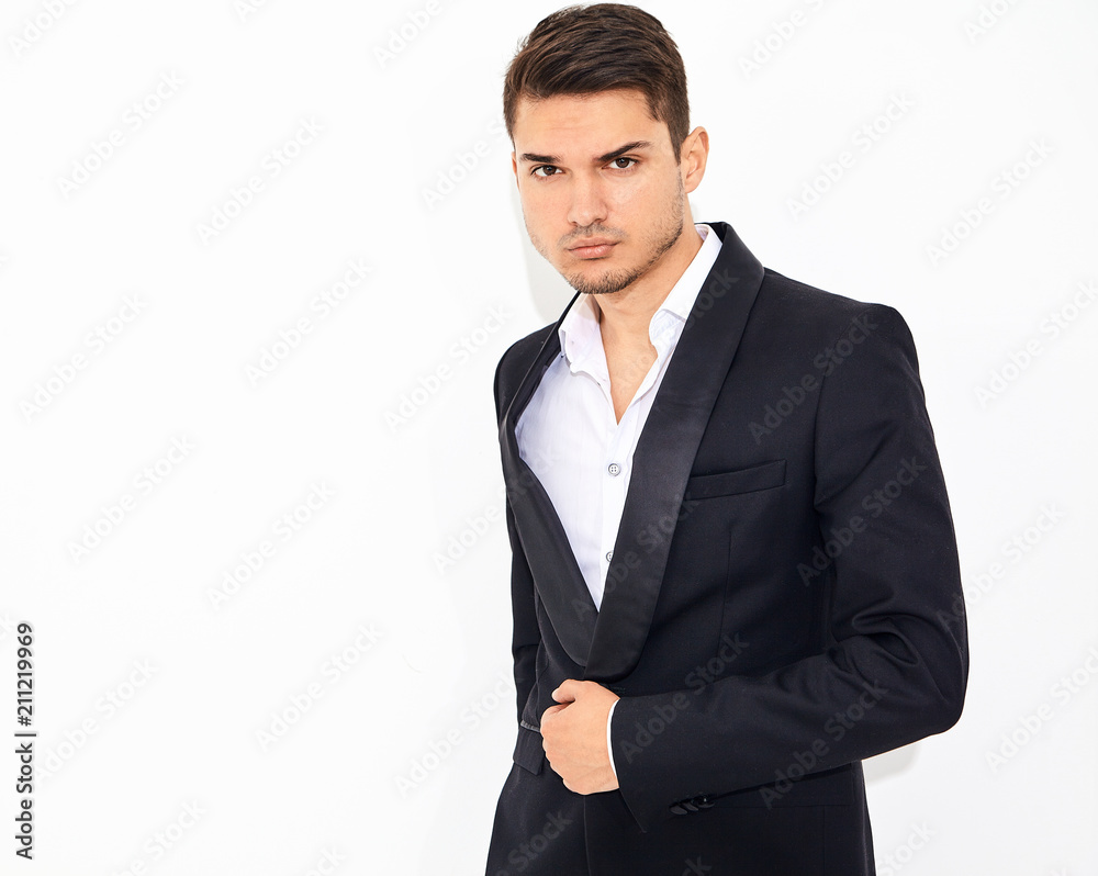 portrait of handsome fashion stylish hipster lumbersexual businessman model dressed in elegant black classic suit posing near white wall in studio. Metrosexual