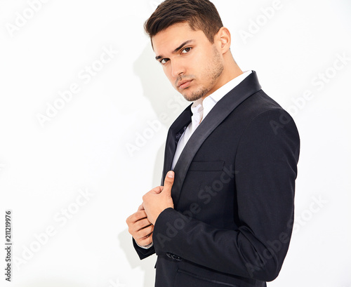 portrait of handsome fashion stylish hipster lumbersexual businessman model dressed in elegant black classic suit posing near white wall in studio. Metrosexual