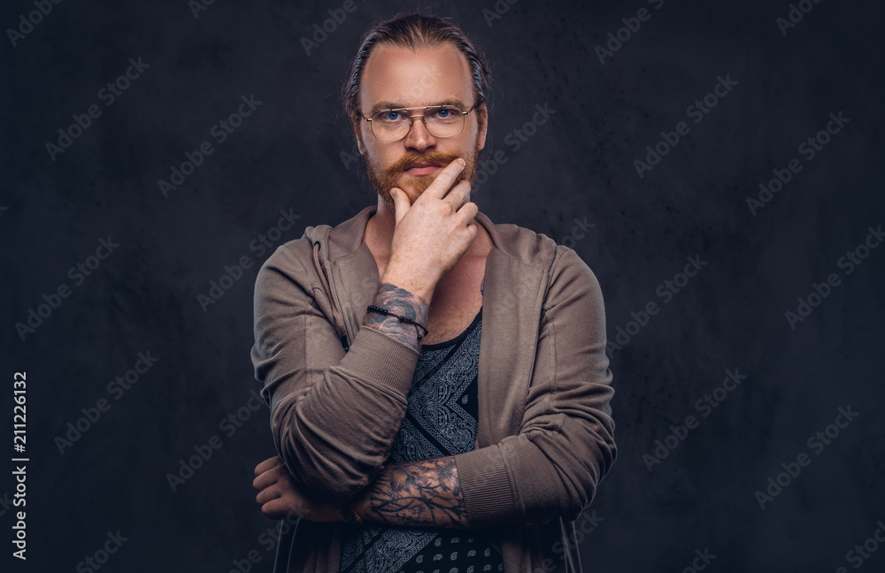 Smart pensive redhead hipster with full beard and glasses dressed in casual clothes, poses with hand on chin in a studio. Isolated on a dark background. Smart pensive redhead hipster with full beard and glasses dressed in casual clothes, poses with hand on chin in a studio. Isolated on a dark background.
