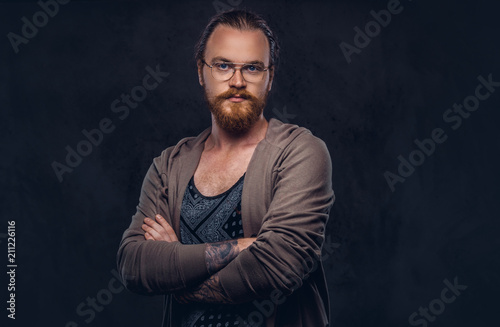 Portrait of a redhead hipster male dressed in casual clothes with glasses and full beard, standing with crossed arms in a studio. Isolated on the dark background.