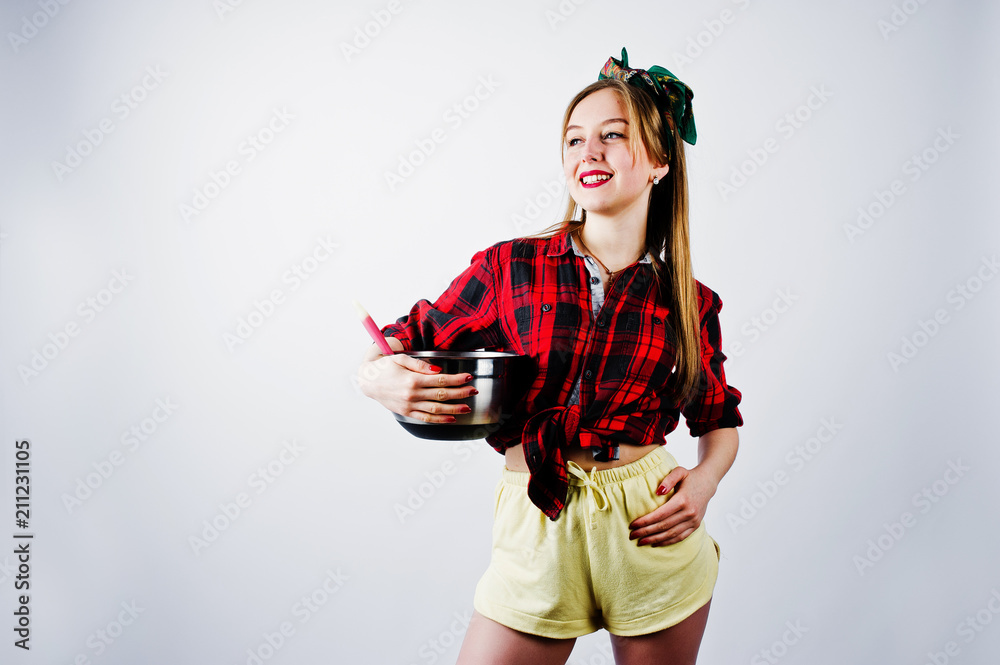 Young funny housewife in checkered shirt and yellow shorts pin up style with saucepan and kitchen spoon isolated on white background.