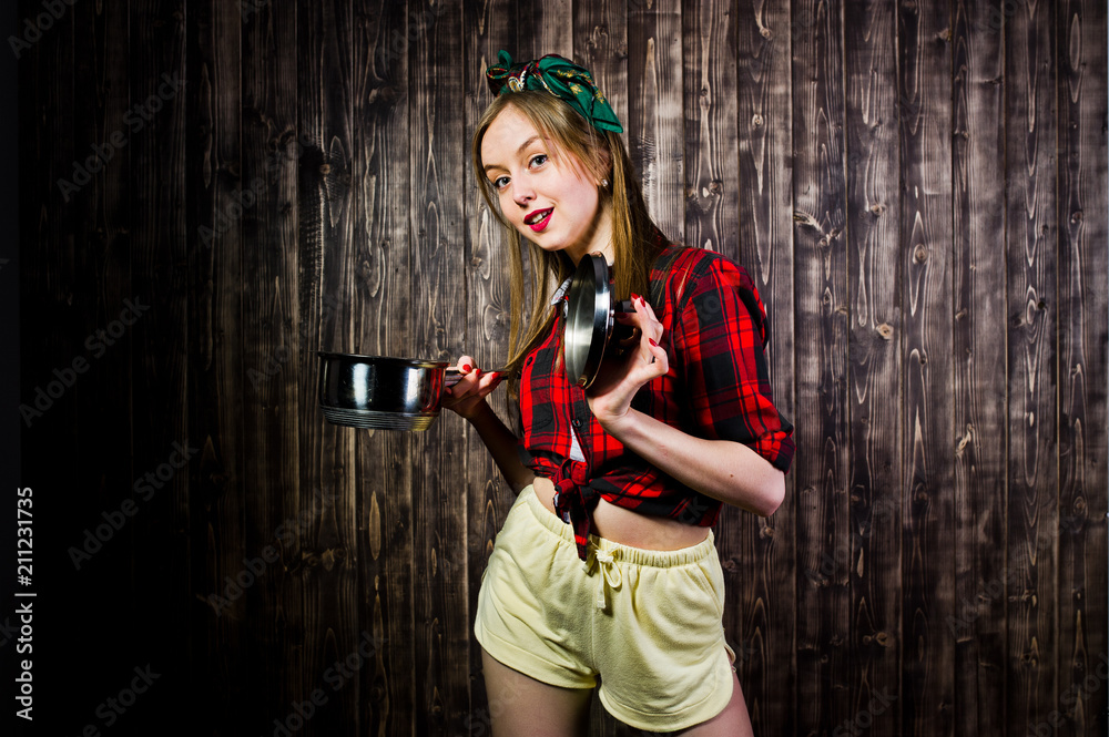 Fototapeta premium Young funny housewife in checkered shirt and yellow shorts pin up style with saucepan on wooden background.