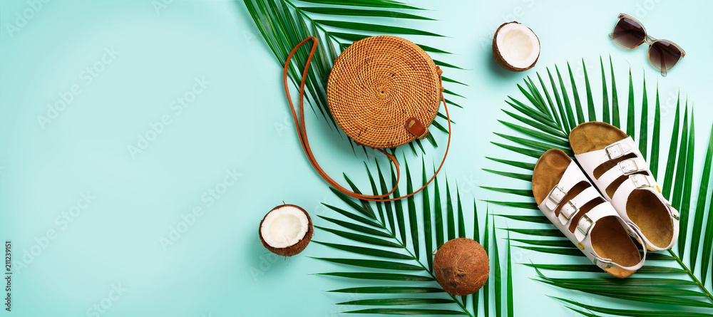 Round rattan bag, coconut, birkenstocks, palm branches, sunglasses on blue background. Banner. Top view with copy space. Trendy bamboo bag and shoes. Summer fashion flat lay. Trip, vacation concept - obrazy, fototapety, plakaty 