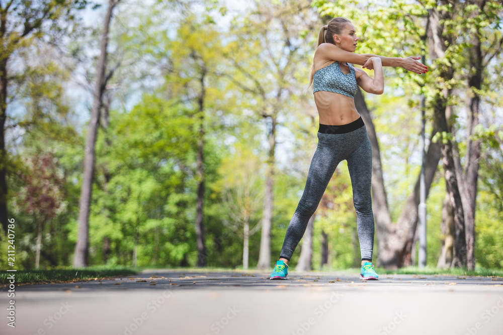 Athletic woman is recharging from nature while training. She is standing on path and stretching arm. Copy space in left side