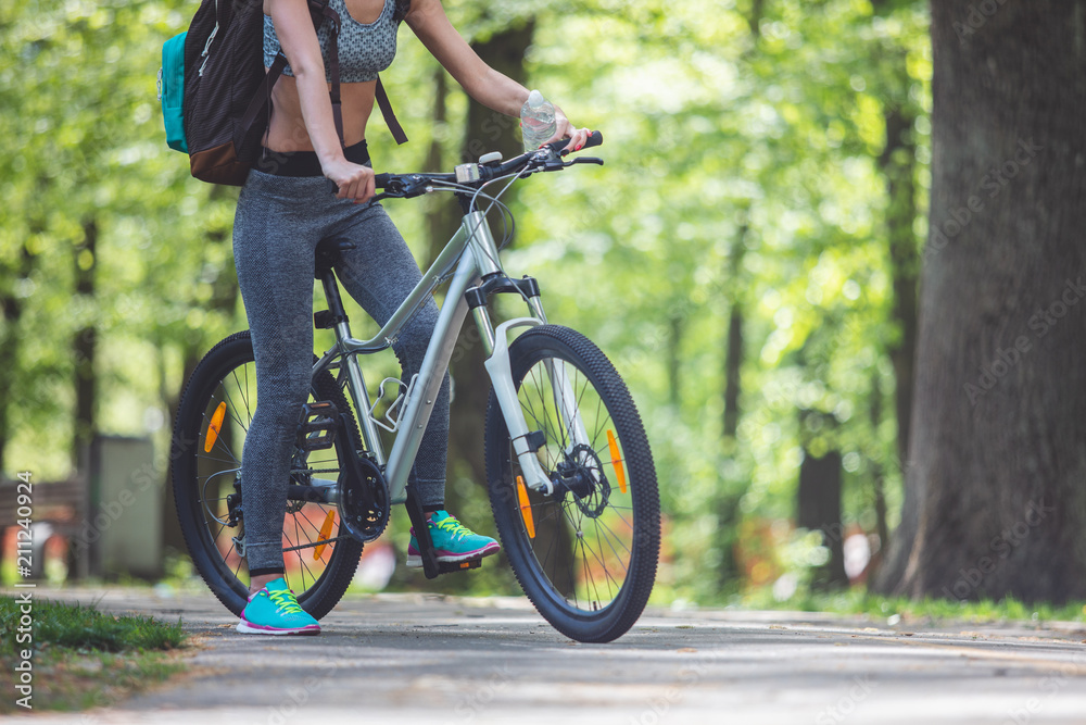Fototapeta premium Low angle of sporty lady riding bike among green landscapes. She is carrying backpack and holding bottle of water. Travelling on bicycle in nature concept