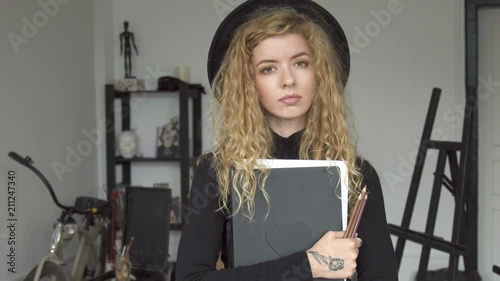 Portrait of young female artist, beautiful girl with curly fair hair standing in the middle of the studio, holding big sketchbook photo