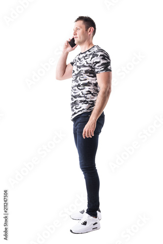 Side view of confident adult man in camo t-shirt talking on the mobile phone and looking away. Full body isolated on white background. 