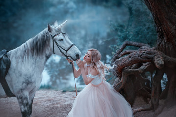 A fairy in a tender vintage dress hugs a unicorn. Fantastic magical, radiant horse. Background river and forest. Blonde girl with wavy hair - light elf. Artistic Photo