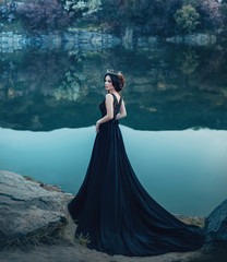 A majestic lady, a dark queen, stands on the background of a river and rocks, in a long black dress. The brunette girl in the gothic crown. Gloomy tone, artistic retouching