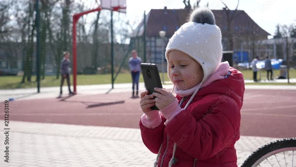 Child girl watch smart phone outdoors while other children play on sports playground Child girl watch smart phone outdoors while other children play on sports playground