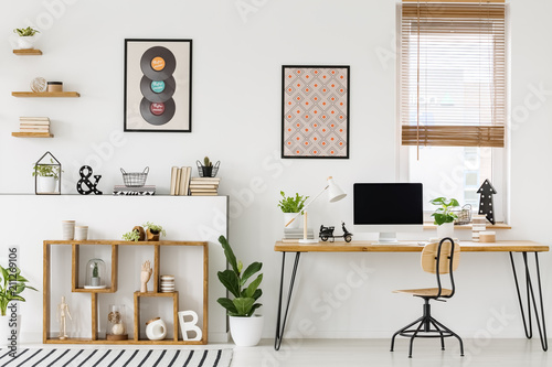 Real photo of a desk with a computer screen standing with a chair next to shelves with ornaments in a bright workplace interior with posters on a wall and window with blinds © Photographee.eu