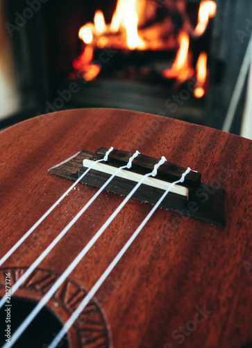 Ukulele small guitar close up stings, fireplace on the background. Musical concept, guitar fret board macro, fire in chimney, cosy romantic atmosphere.