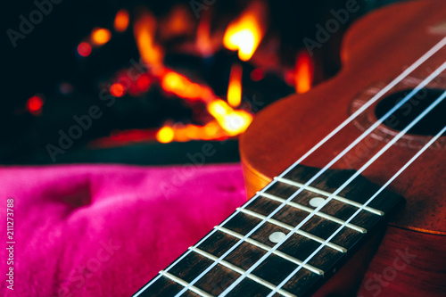 Ukulele small guitar close up stings, fireplace on the background. Musical concept, guitar fret board macro, fire in chimney, cosy romantic atmosphere.